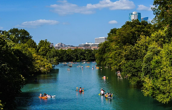 People kayaking and paddleboarding on a tree-lined urban lake, with a city skyline in the background.