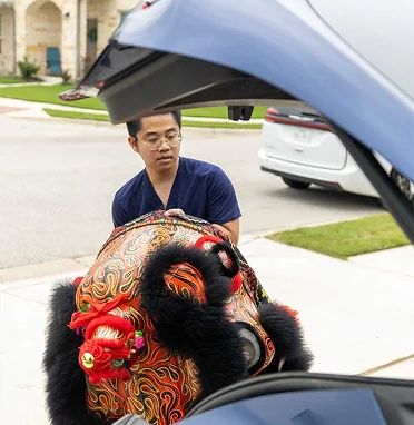 Kevin Glan carefully places a large Lion Dance head into the trunk of a car, showing the blend of ancient tradition and modern life.