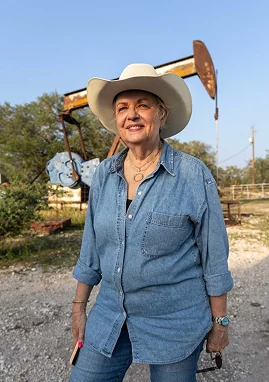 Marsha Hendler in a cowboy hat stands proudly in front of an oil derrick, defying expectations of a typical oiler.