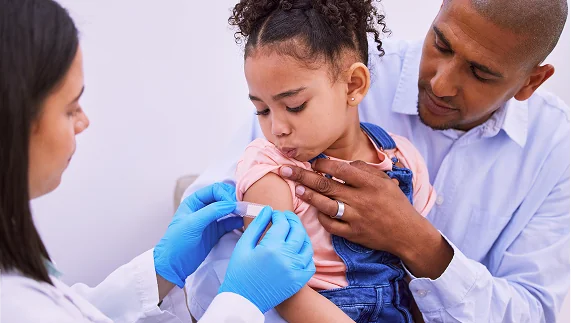 A young girl bravely receives a flu shot from a doctor while her father comforts her, showing proactive health measures.