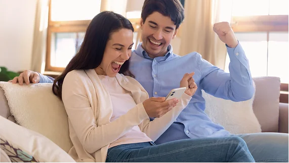A young couple on a couch celebrates with excitement as they look at a smartphone, pleased with the TxT app.